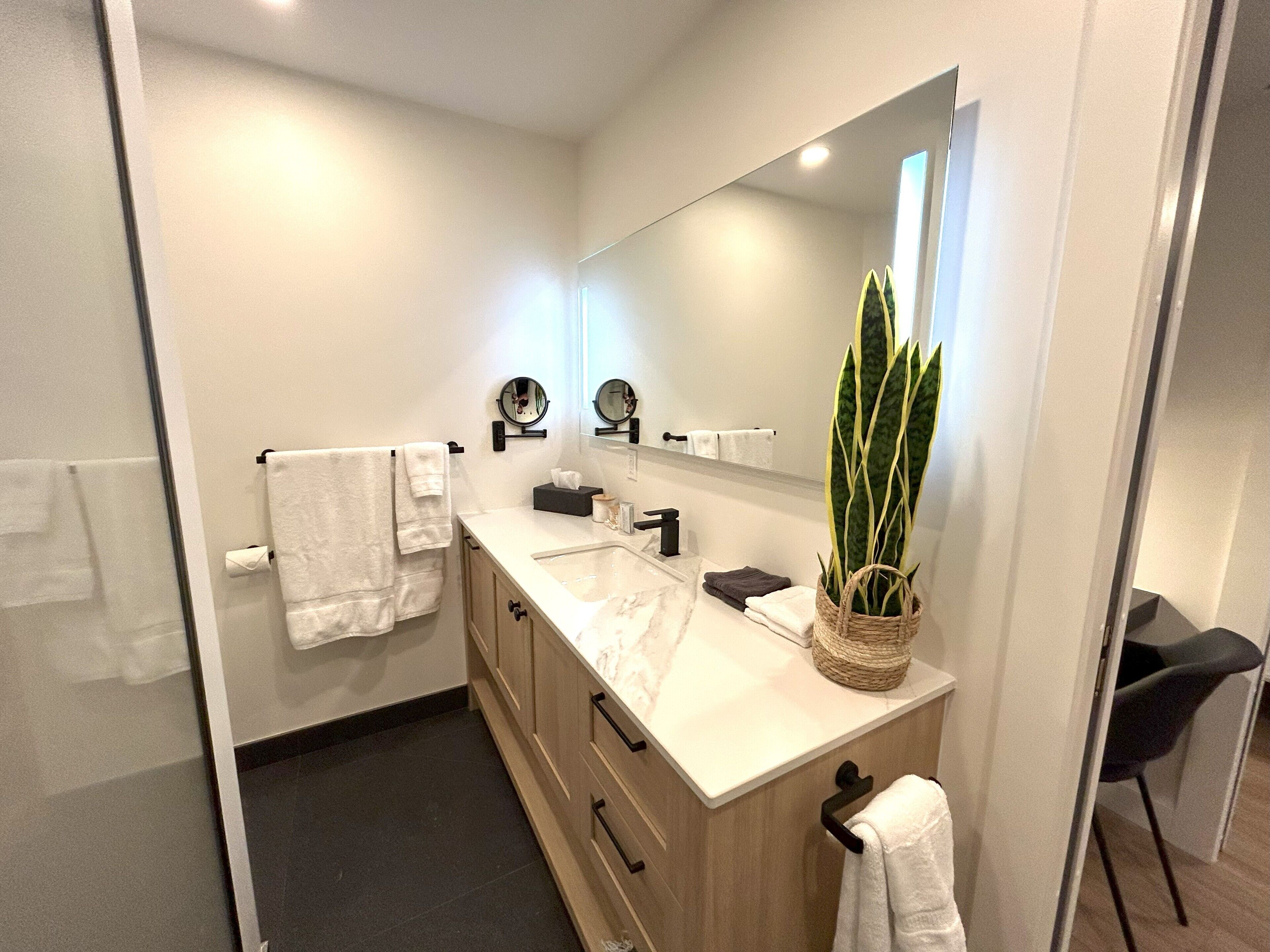 A modern bathroom with a large mirror, a sink, and a vanity. There are multiple towels hanging on hooks, a potted plant on the vanity, and a small table with a basket.
