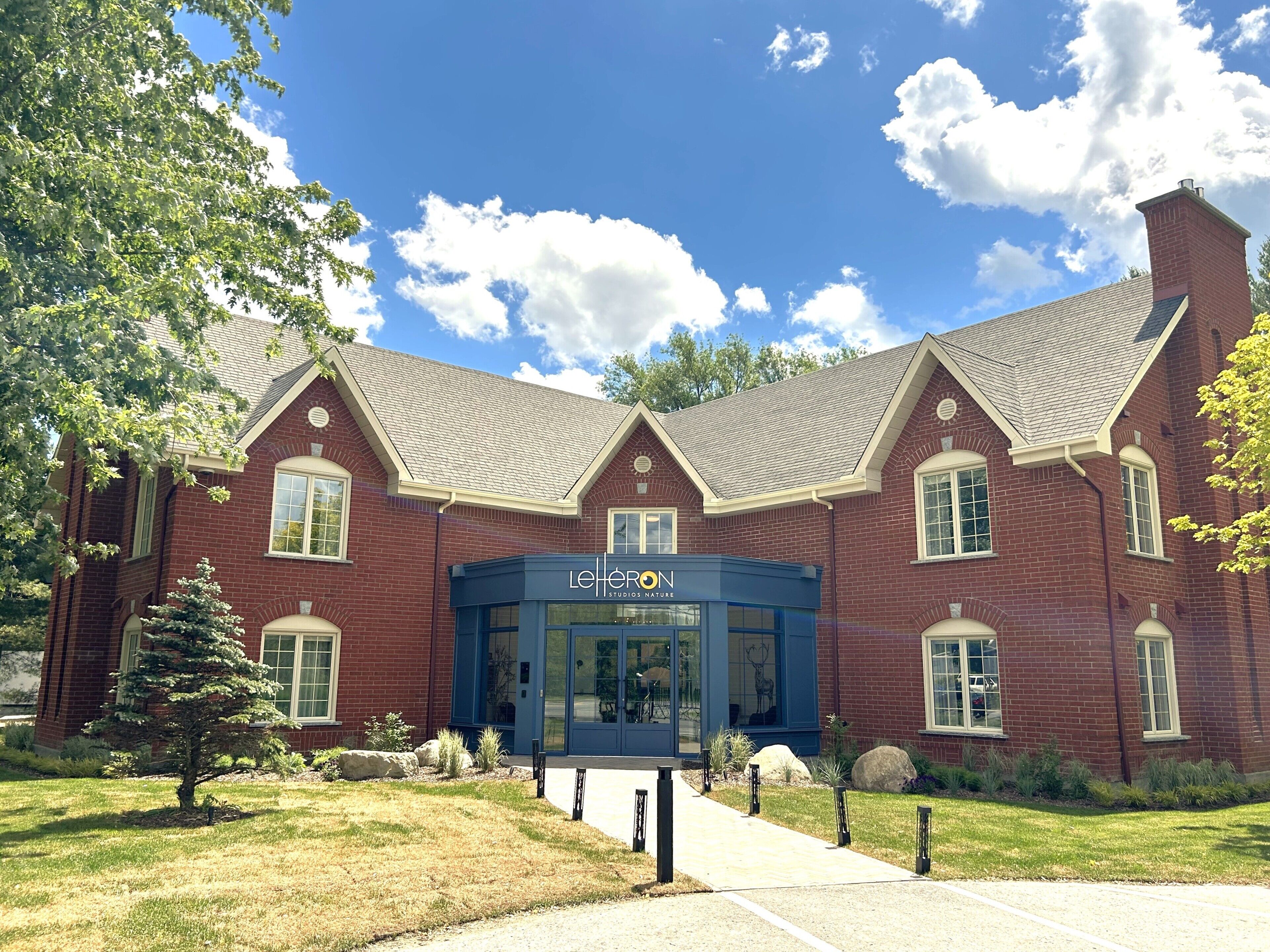 A brick building with a blue entrance, a sign reading "LeHuron Studios Nature," and a well-maintained lawn with a small tree.
