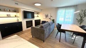A modern living room with a fireplace, a sofa, a dining table, and a TV mounted on the wall. - Le Héron - Studios Nature (Magog)