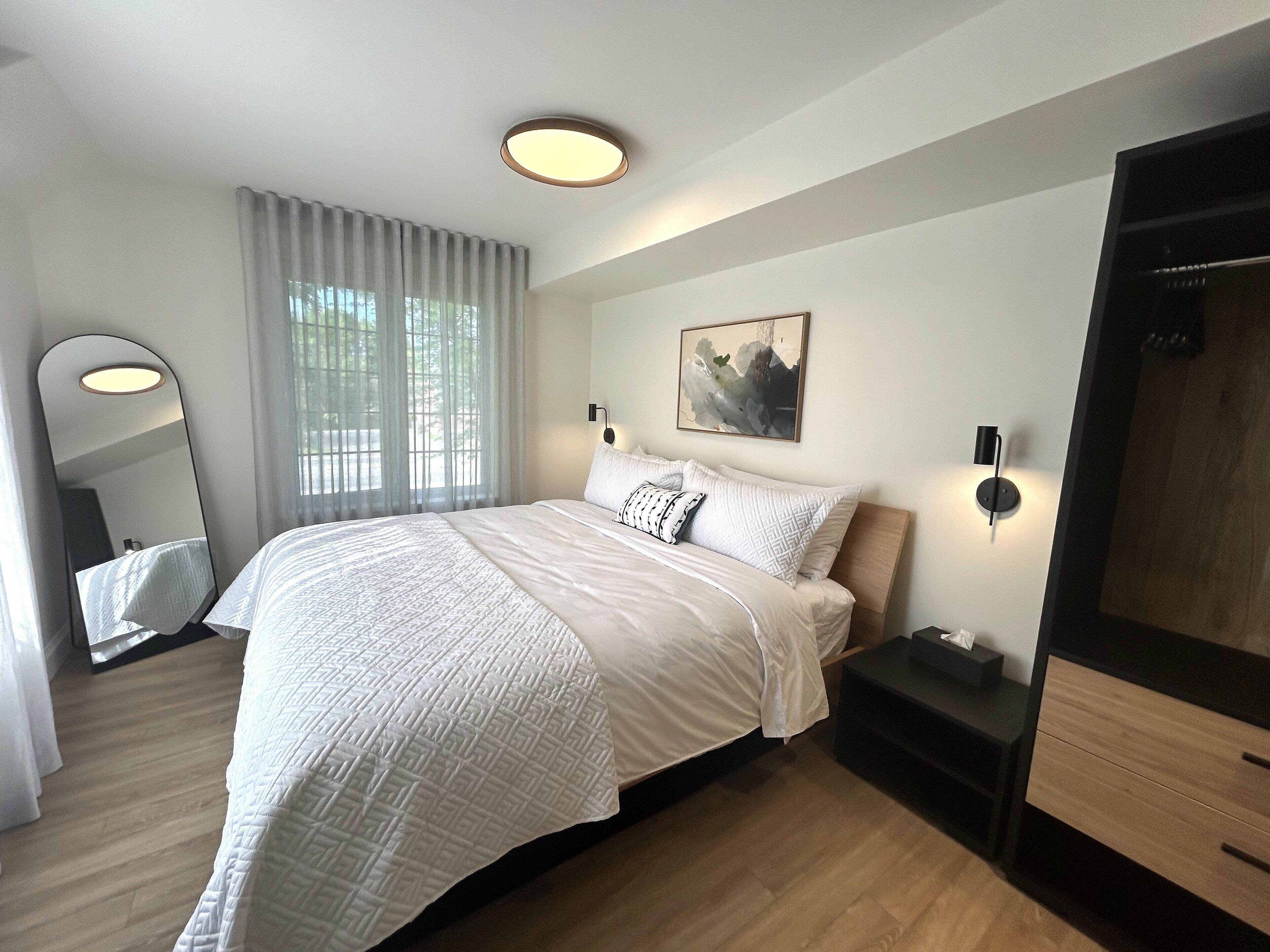 A modern bedroom with a large bed, a bedside table, a mirror, and a wall-mounted light fixture.