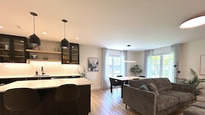 A modern kitchen with a central island, dining area with a table and chairs, and a living room with a sofa and a plant. - Le Héron - Studios Nature (Magog)