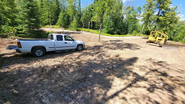 Un camionnette blanche était garée sur un chemin de terre, dans une zone boisée, à proximité d’un engin de chantier jaune.