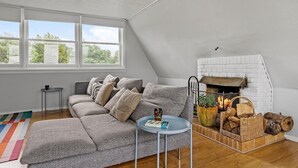 Una sala de estar acogedora con un sofá modular, una mesita redonda con un libro, una chimenea de ladrillo blanco y un ventanal con vistas a árboles.