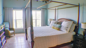 A four-poster bed with a quilt, a bedside table with a lamp, and a chair in a room with a large window and curtains. - NEW LISTING!! The ULTIMATE Sunrise Retreat! (St. John)
