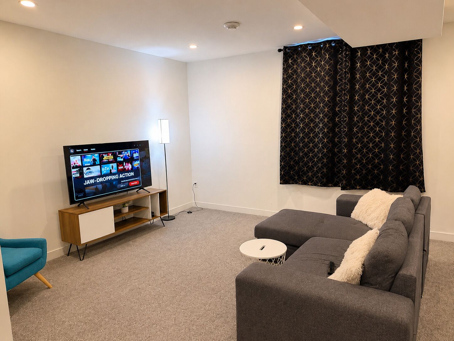 Una sala de estar con un televisor de pantalla plana, un sofá gris, una mesita redonda y una lámpara de pie.
