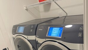 Vista frontal de uma máquina de lavar e secar Samsung, ambas com painel digital, instaladas numa lavanderia com chão em mosaico preto e branco.