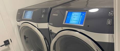 A front view of a Samsung front-loading washer and dryer unit with a digital display, located in a laundry room with a black and white tiled floor.