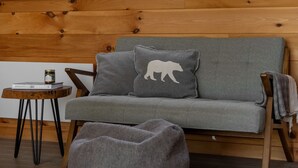 A cozy room with a wooden ceiling, a grey sofa with a bear pillow, a grey bean bag, and a small table with a jar. - Cozy cabin - lake view  (Lac-des-Îles)