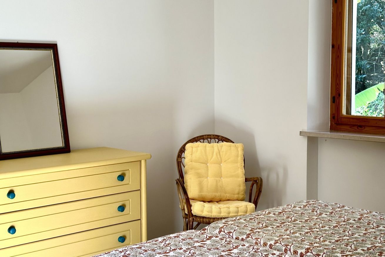 A room with a bed, a yellow dresser with drawers, a wicker chair with a yellow cushion, a mirror on the dresser, and a window with a view of greenery.