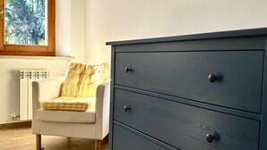 Una habitació amb una cadira blanca, un calaixera fosc amb calaixos, un radiador i una finestra amb vista a arbres.