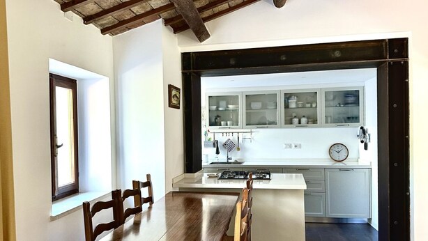A dining area with a wooden table and chairs, a kitchen with cabinets and a clock, and a window with a door. - S333 – Sirolo, three-room apartment with terrace and parking space (Sirolo)
