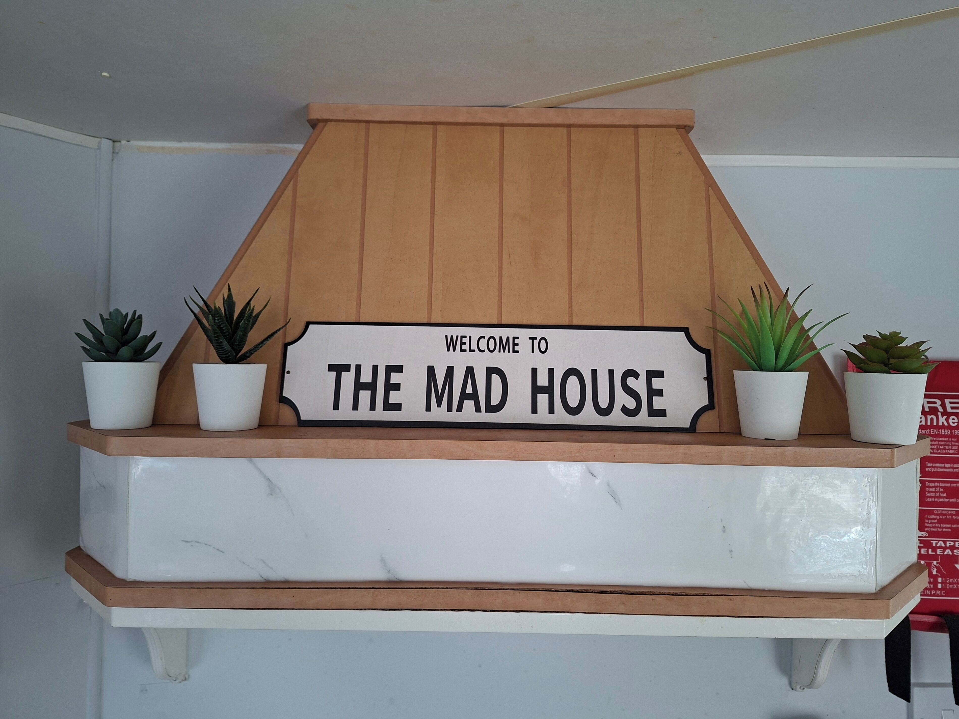 A welcome sign for 'THE MAD HOUSE' above a marble shelf with potted plants.