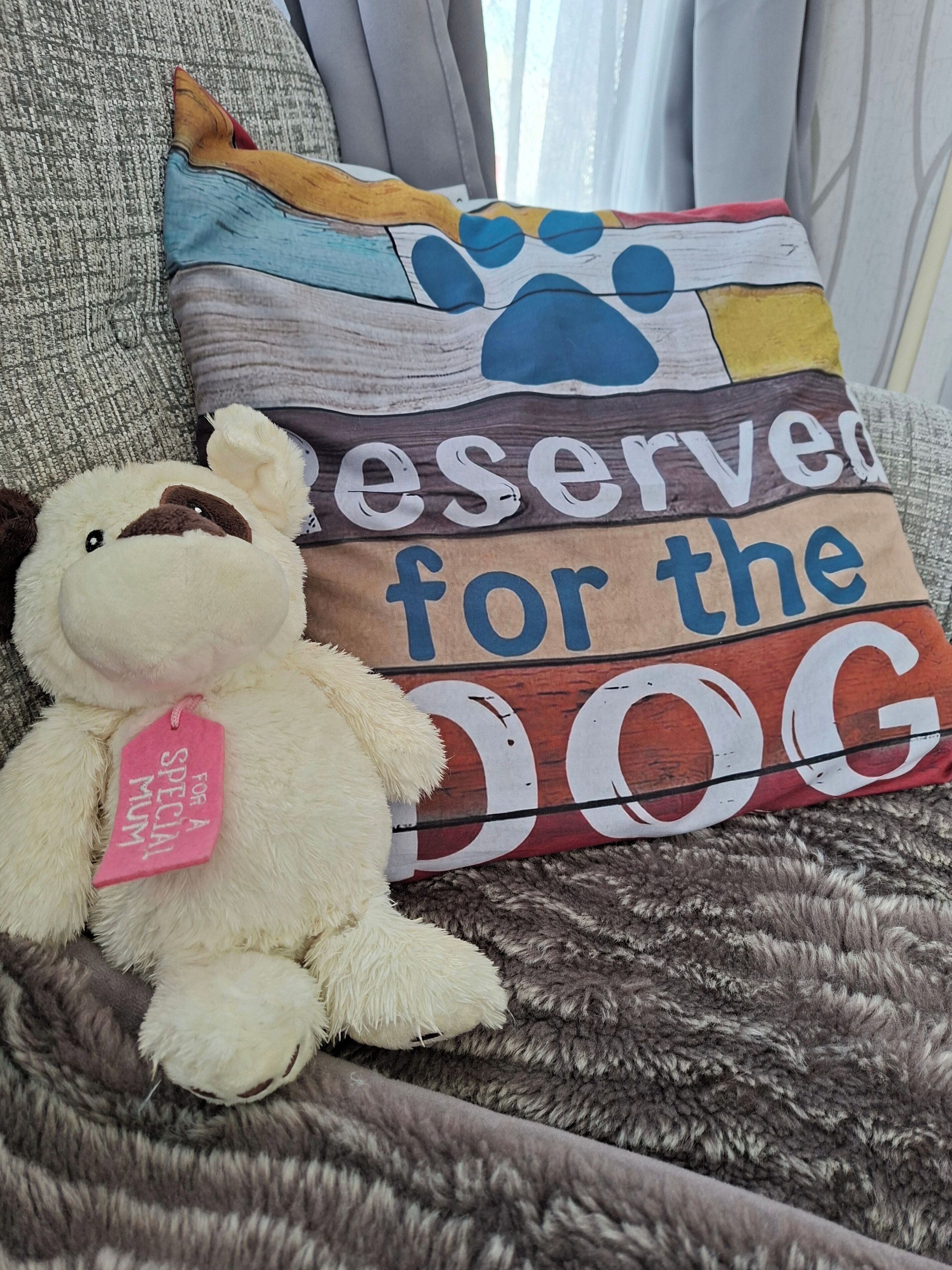 A plush toy with a tag on a textured fabric surface, possibly a couch or bed, with a pillow featuring a dog-themed design.