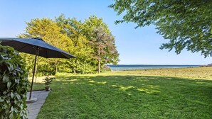 Ein grüner Außenbereich mit einer Terrassentischgarnitur und Sonnenschirm, Bäumen und Blick auf das Meer.