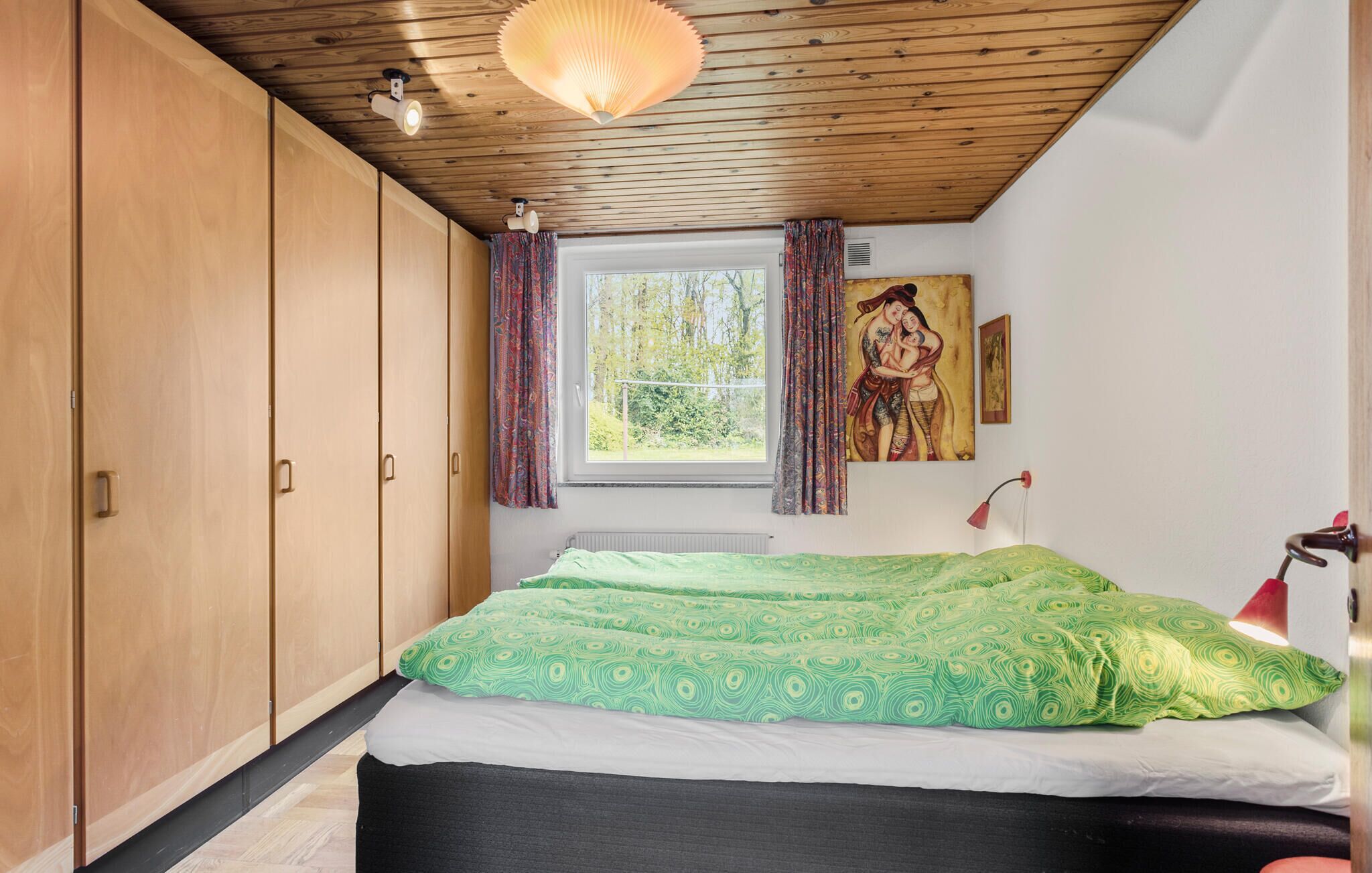A bedroom with a large bed, wooden wardrobe, a window with curtains, a painting on the wall, and a ceiling light.