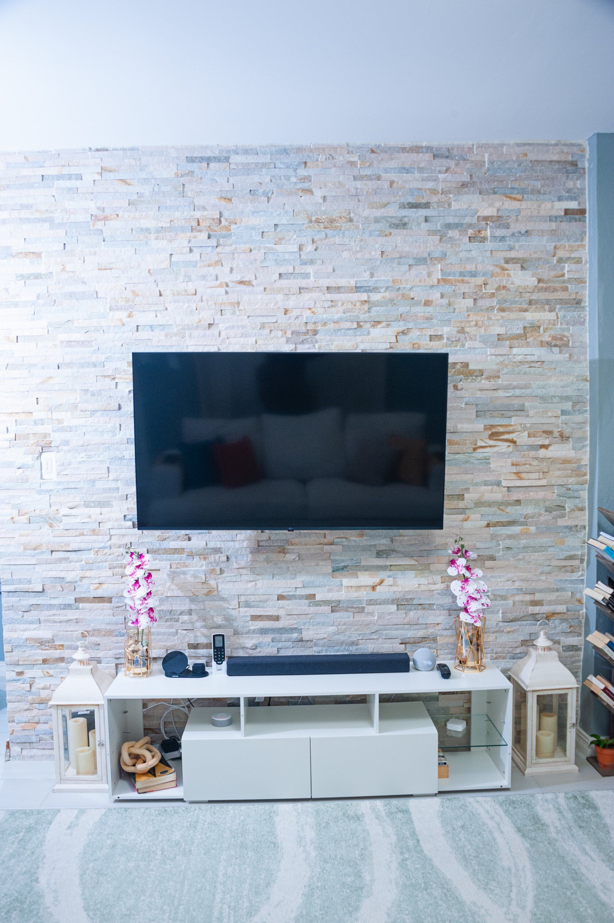 A modern living room with a stone wall, a flat-screen TV mounted above a white entertainment unit, and decorative items like a lantern and flowers.