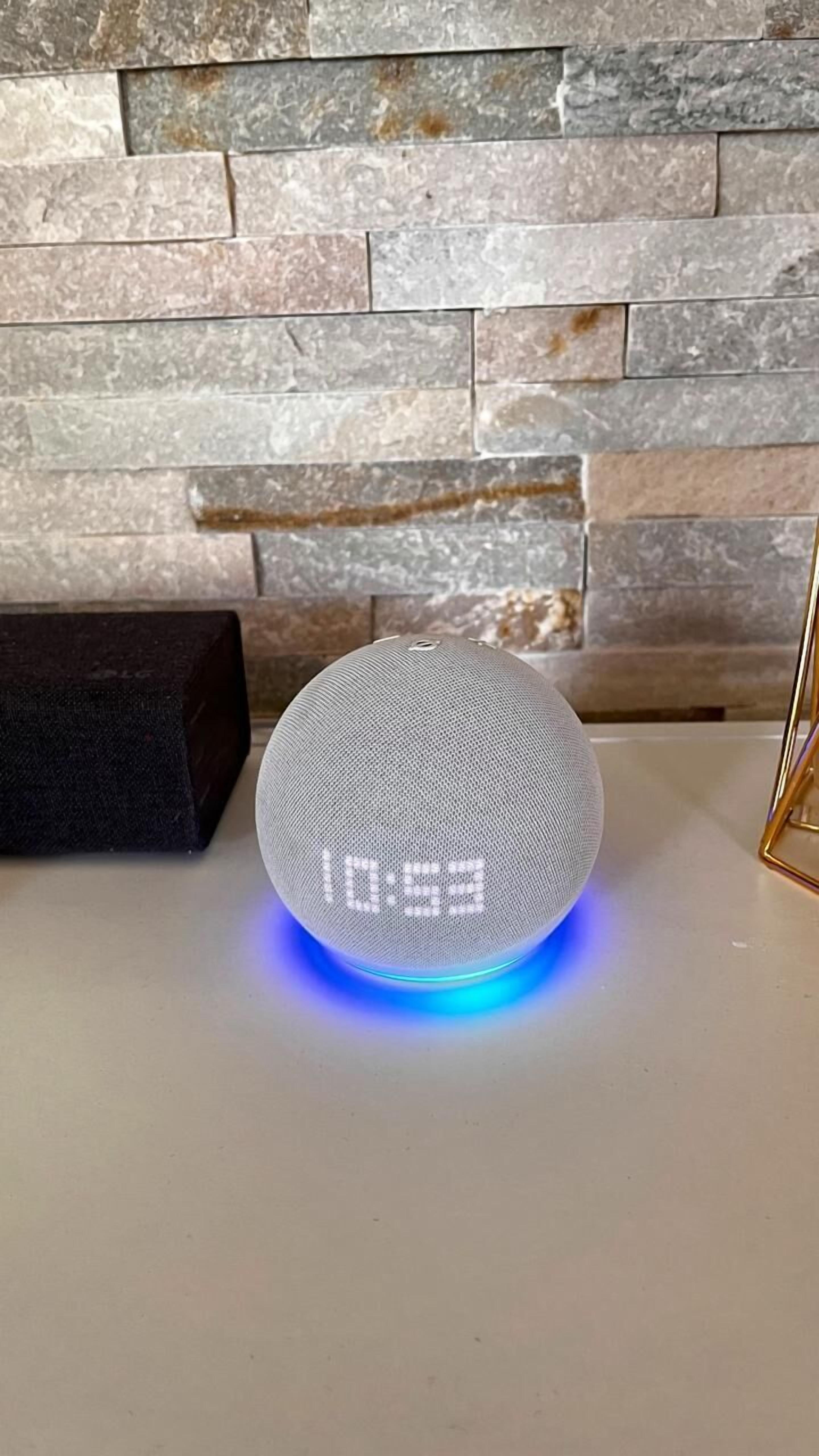 Un altavoz redondo y blanco con pantalla digital sobre una superficie blanca, con un fondo de pared de piedra.
