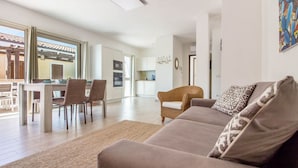 A living room with a sofa, chairs, a dining table, and a kitchen in the background. - GuestHost - Brand New Apartment with Pool and Terrace - Two Steps from the Beaches (Palau)