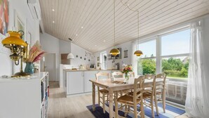 A modern kitchen with white cabinets, a dining area with a wooden table and chairs, and large windows offering a view of greenery. - Amazing home in Tarm with sauna (Tarm)