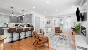 A modern living room with a sofa, armchairs, and a coffee table. There is a kitchen area with bar stools and a dining table. - Home Away From Home in Lowell (Lowell)