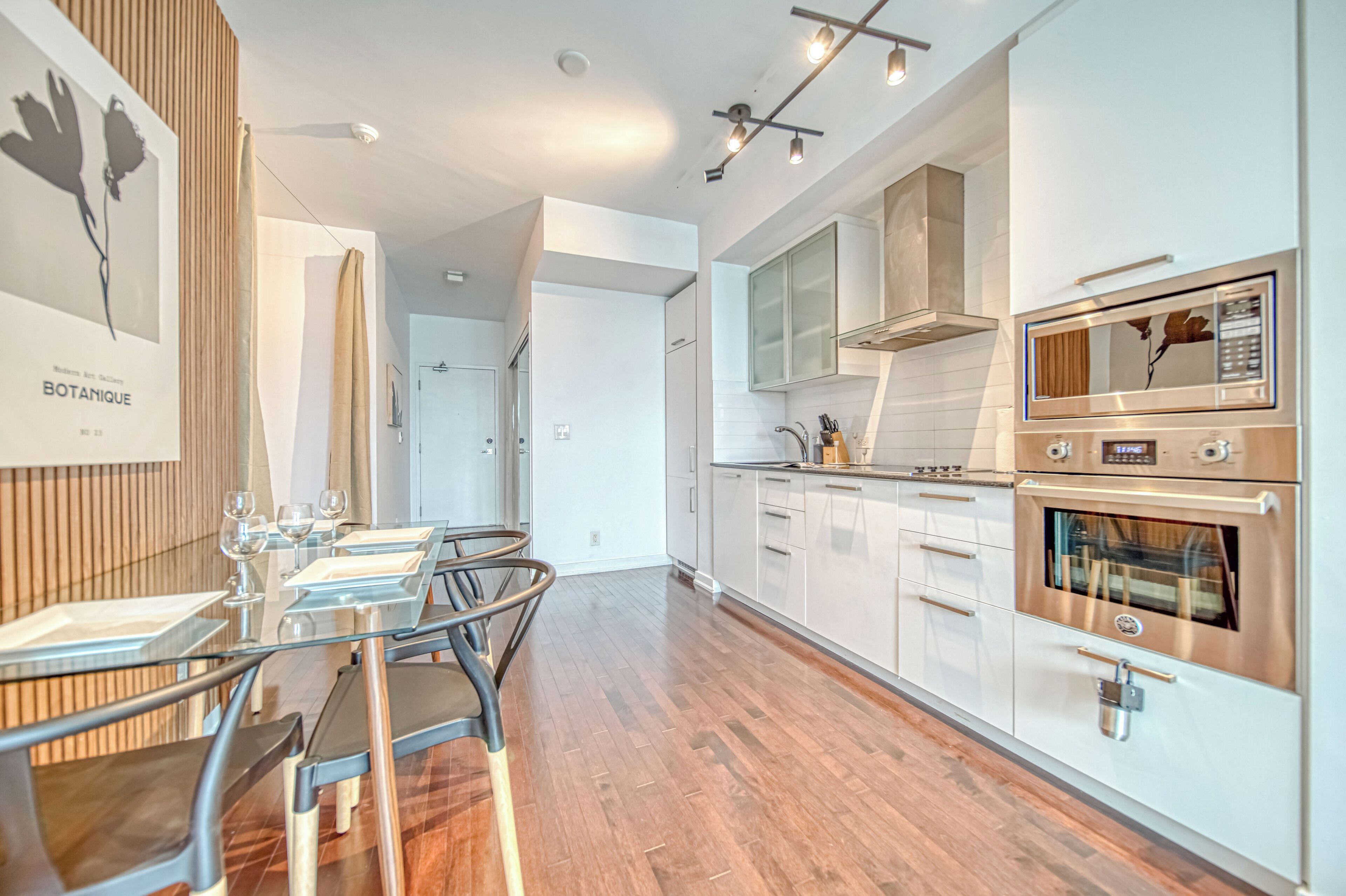 Una cocina moderna con armarios blancos, electrodomésticos de acero inoxidable y una zona de comedor con mesas y sillas de vidrio.