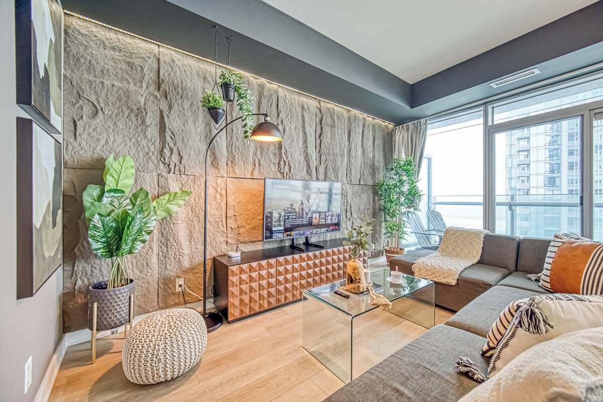 A modern living room with a stone accent wall, a flat-screen TV, a sofa, a coffee table, and a large window offering a city view.