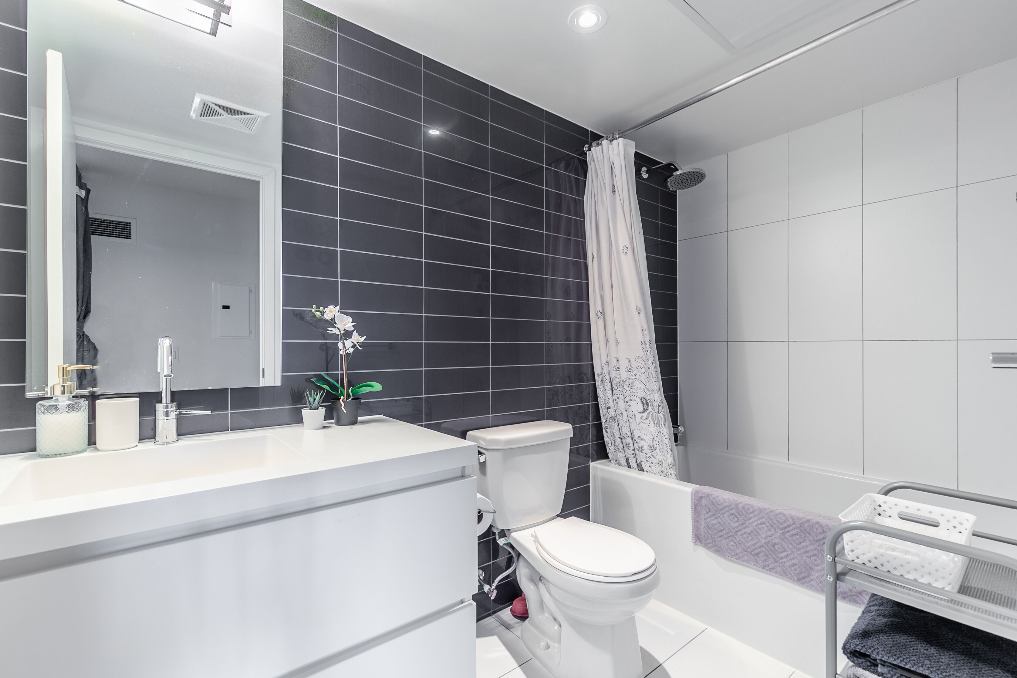 A modern bathroom with a white sink, toilet, and bathtub. There is a shower curtain, a towel rack, and a potted plant on the counter.