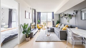 A modern living room with a grey sofa, yellow chair, and a white desk. There are plants, a TV, and a large window with curtains. - SkyTree Modern Suites (Toronto)