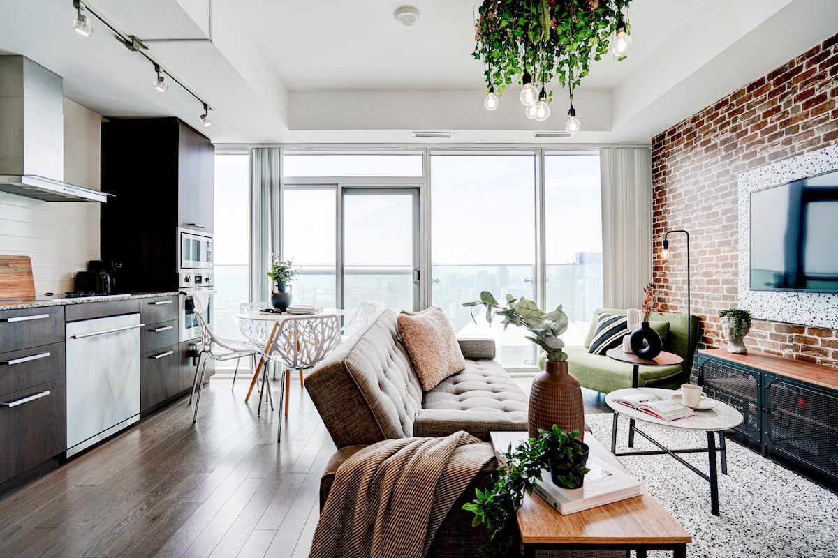 A modern apartment with a kitchen, dining area, and living room. It features large windows, a brick wall, and a hanging plant.