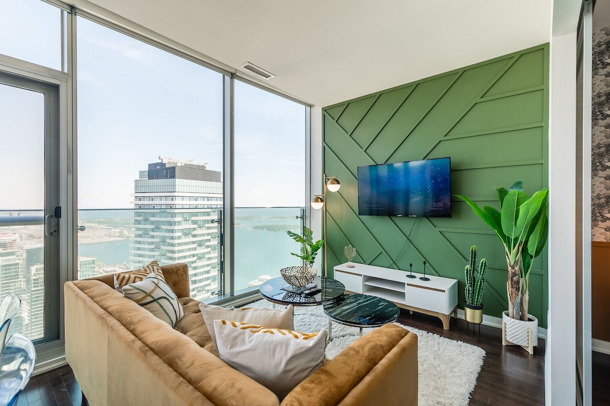 A modern living room with a large window, a green accent wall, a flat-screen TV, and a view of a cityscape.