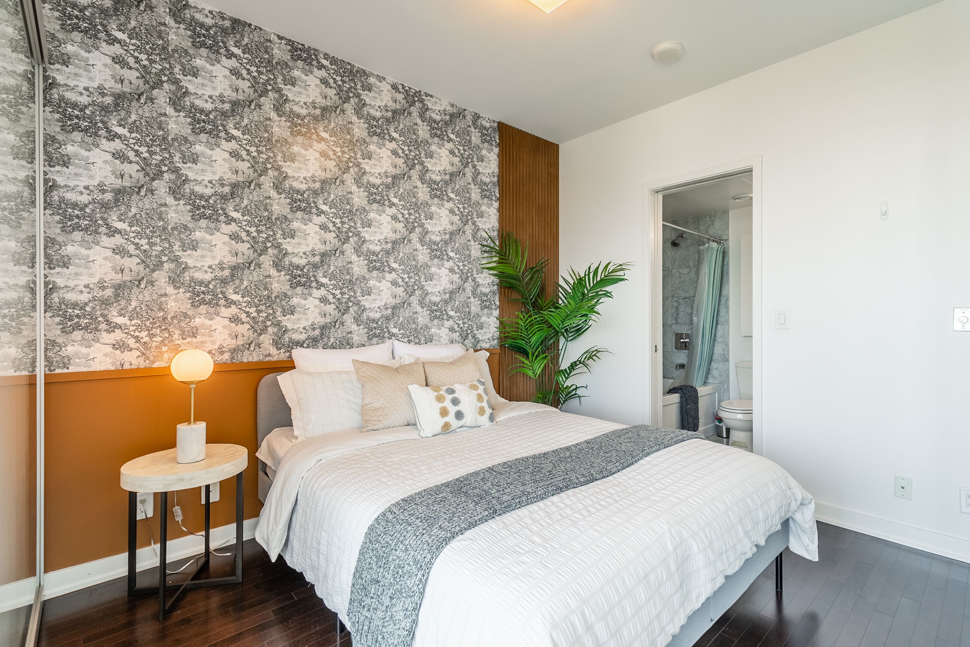 Een moderne slaapkamer met een groot bed, een rond nachtkastje met lamp, een muur met een structuur en een plant.