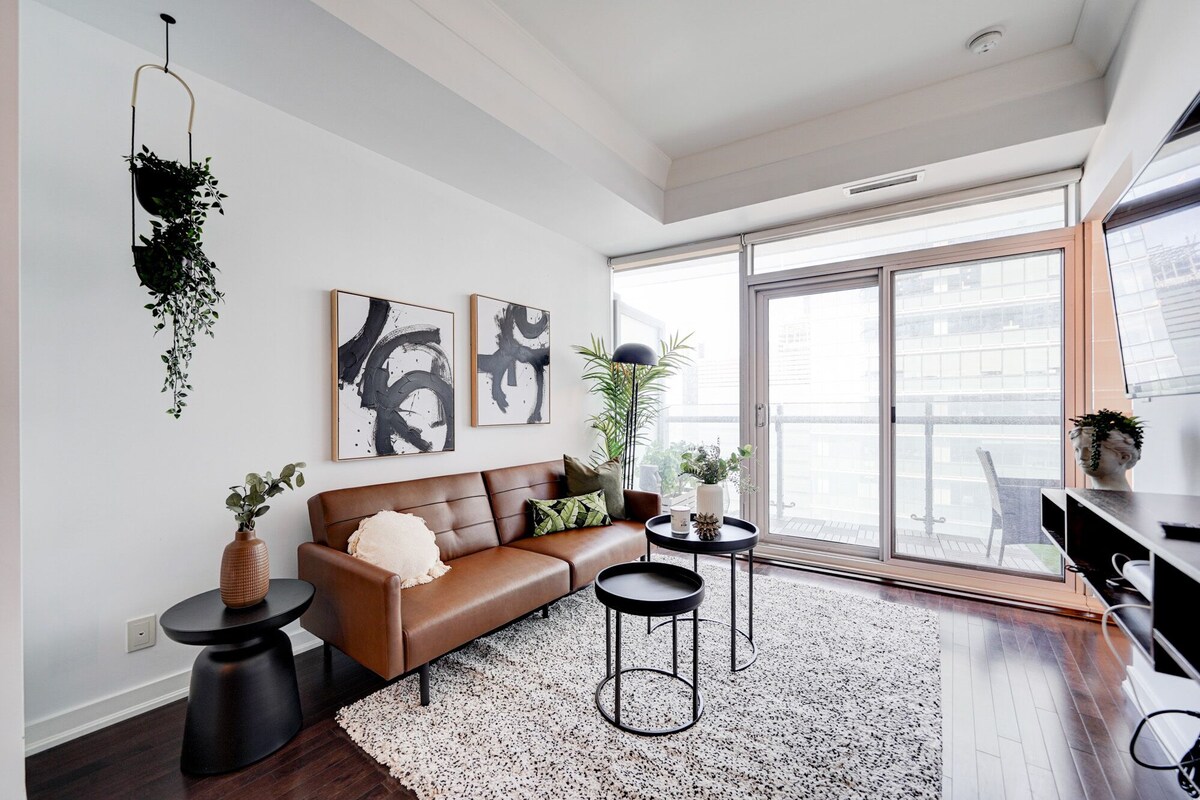 A modern living room with a brown leather sofa, two black side tables, and a black and white rug. There are abstract paintings on the wall, a hanging plant, and a large window with a view of buildings.