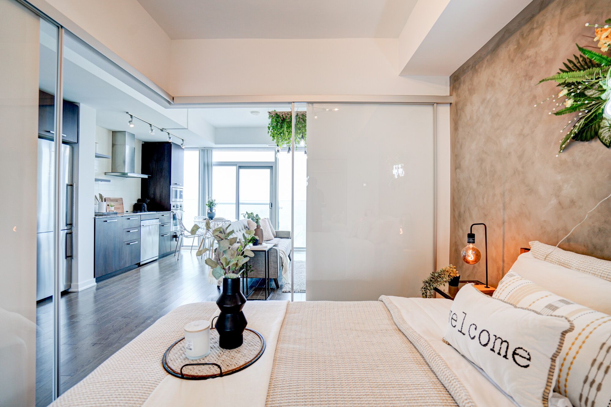 A modern bedroom with a large bed, a bedside table with a lamp, and a view into the kitchen and living area.