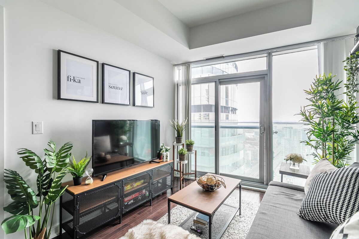 A modern living room with a large TV, a sofa, and a dining table. There are potted plants and a view of the city through large windows.