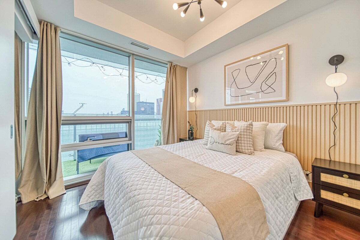A modern bedroom with a large bed, a view of the city, and a framed abstract artwork on the wall.