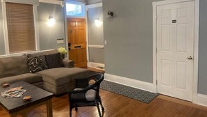 A living room with a grey sofa, a coffee table, and a wicker chair. There is a wooden door and a white door in the background. - Beautiful High-End 4 Bed 3 Bath House, Sleeps 15! (Pittsburgh)