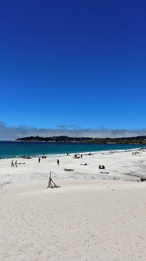 A wide sandy beach with clear blue water and a distant tree-covered hill.