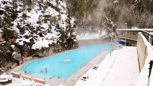 Una piscina esterna coperta di neve con persone a nuoto, circondata da alberi innevati e una scogliera rocciosa.