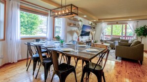Zona pranzo con tavolo in legno apparecchiata per quattro persone, circondata da sedie, e un angolo salotto con divano e televisione.