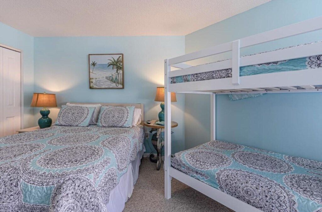 Un dormitorio con una cama litera, una cama individual, ropa de cama estampada, dos lámparas y un cuadro con motivos playeros en la pared.