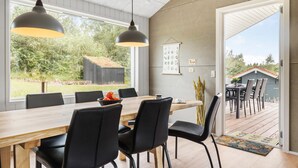 A dining area with a long wooden table, black chairs, and pendant lighting. There is a view of a garden and a wooden structure outside.