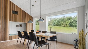 A modern dining area with a wooden panel wall, a large window with a view of greenery, and a table set with chairs and a vase of flowers.