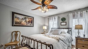 A bedroom with a bed, a chair, a nightstand, a lamp, a ceiling fan, a framed picture, and a window with curtains. - Welcome to our lake house on the beautiful Hiwassee River! (Calhoun)