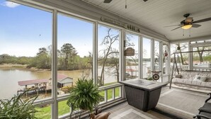 A covered porch with large windows overlooking a lake, a wicker chair, and a table with a plant. - Welcome to our lake house on the beautiful Hiwassee River! (Calhoun)