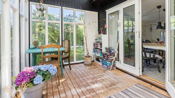 Eine gemütliche Terrasse mit Holzboden, einem Esstisch mit Stühlen, einem Blumentopf und einer Glastür zu einem Innenraum.