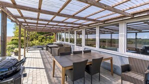 A covered patio area with a wooden pergola, outdoor seating, and large windows offering views of the surrounding landscape. - 3 bedroom beautiful home in Rønne (Rønne)
