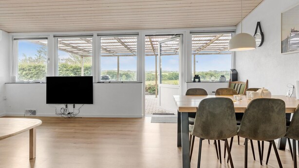 A spacious dining area with a large table and chairs, a flat-screen TV mounted on the wall, and large windows offering a view of the outdoors. - 3 bedroom beautiful home in Rønne (Rønne)