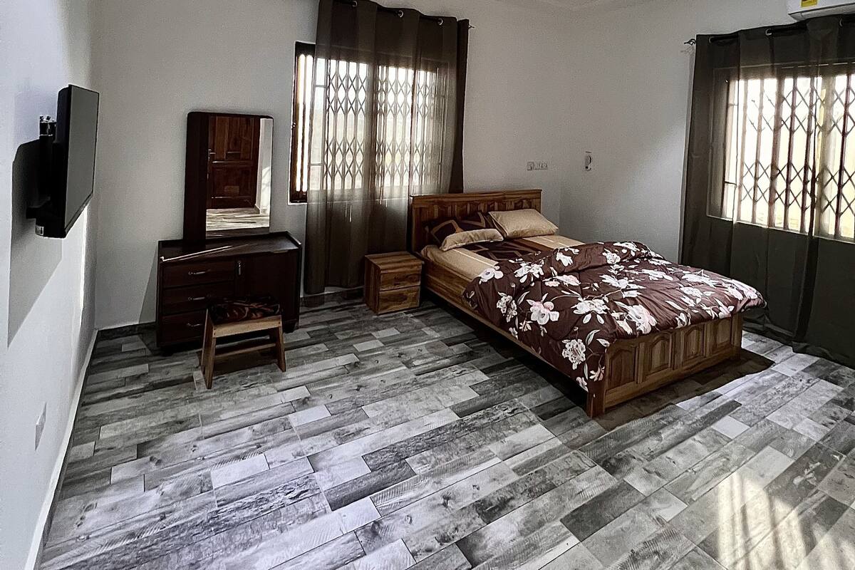 Ein Schlafzimmer mit einem Bett, einem Schminktischchen, einem Kleiderschrank, einem Deckenventilator und einem an der Wand befestigten Fernseher.