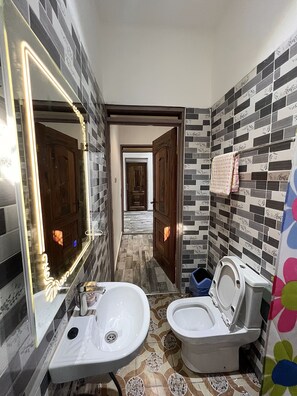 Un baño con lavamanos, inodoro y espejo. Las paredes tienen azulejos estampados, y hay una puerta que conduce a otra habitación.