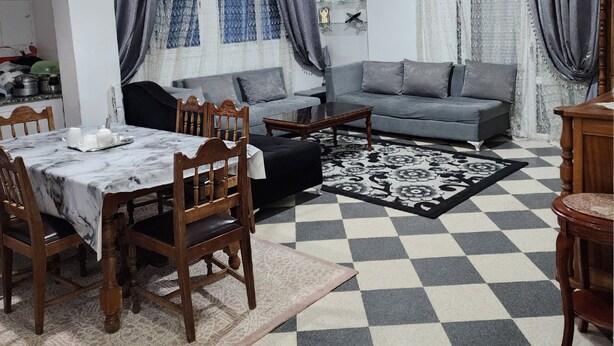 Una sala de estar con piso a cuadros, sofá gris, mesa de comedor con sillas y alfombra estampada.